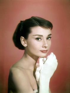 Audrey Hepburn facing the camera against a pink background, her body facing to the right as she rests her chin on her white-gloved hands