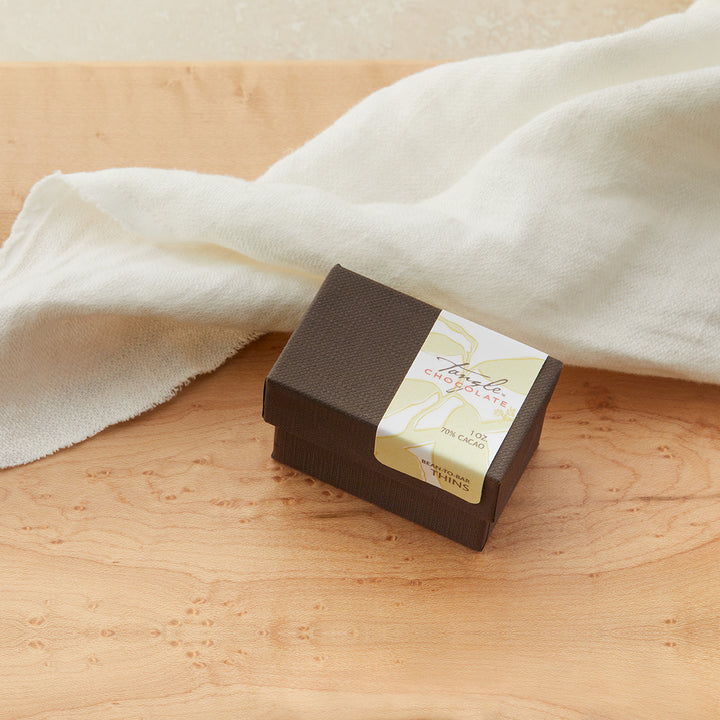 Small closed brown box with white and green label with leaves that says Tangle Chocolate. On a maple table with a white cloth draped behind it.