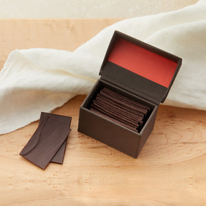 Small open brown box with coral color inside the lid. Filled with slivers of dark chocolate with a couple of the slivers to the side of the box. On a maple table with a white cloth draped behind it.