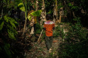 a child in a bright orange patterned shirt, holding a long stick, facing away from the camera, walking amongst the green and brown jungle