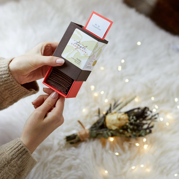 a person sliding open a red and brown box of Tangle Chocolate thins, with small bright lights and a small floral bouquet laying on a white background