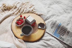 a round wooden breakfast tray containing Tangle Chocolate thins, a cup of coffee, and raspberries on bed with folded newspaper