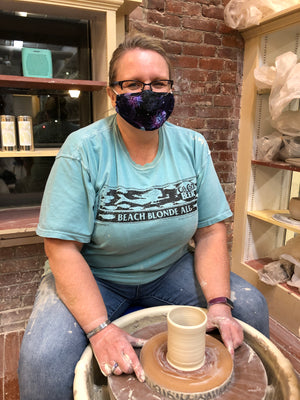 Black Cats Pottery's Helen Thelan wearing a blue t-shirt and jeans, sitting at a pottery wheel, making a hot chocolate mug for Tangle Chocolate