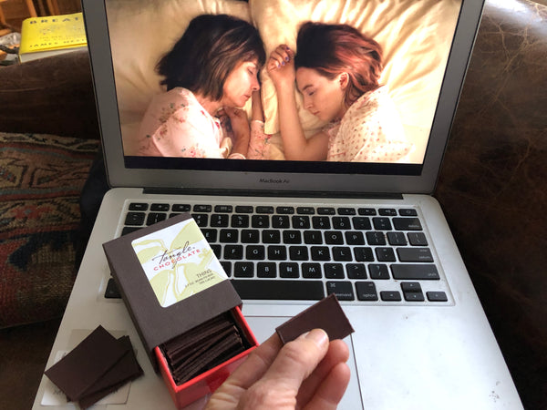 a hand holding a Tangle Chocolate thin in front of a laptop computer monitor, displaying a scene of two women facing one another in bed