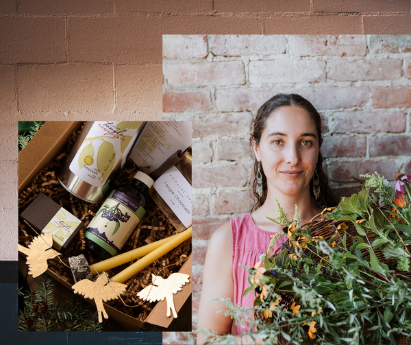 Woman with large basket of herbs and flowers, with gift box with products for the winter: Tangle Hot Chocolate, box of chocolate, soap, candles, elderberry syrup