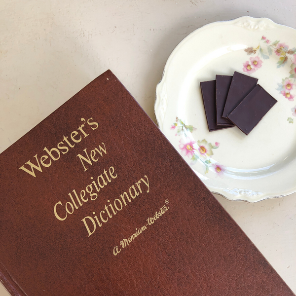 a brown leather book that reads 'WEBSTER'S NEW COLLEGIATE DICTIONARY, MERRIAM WEBSTER' sitting next to a white, round floral dish containing four Tangle Chocolate thins