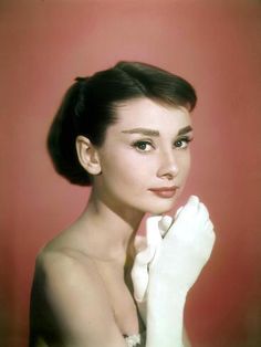 Audrey Hepburn facing the camera against a pink background, her body facing to the right as she rests her chin on her white-gloved hands 