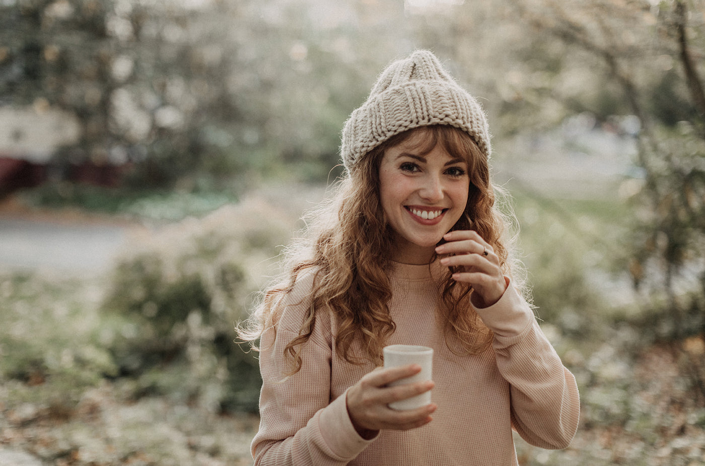 Beautiful white woman with long auburn hair standing outside in pink shirt and winter hat smiling and holding a mug of hot beverage