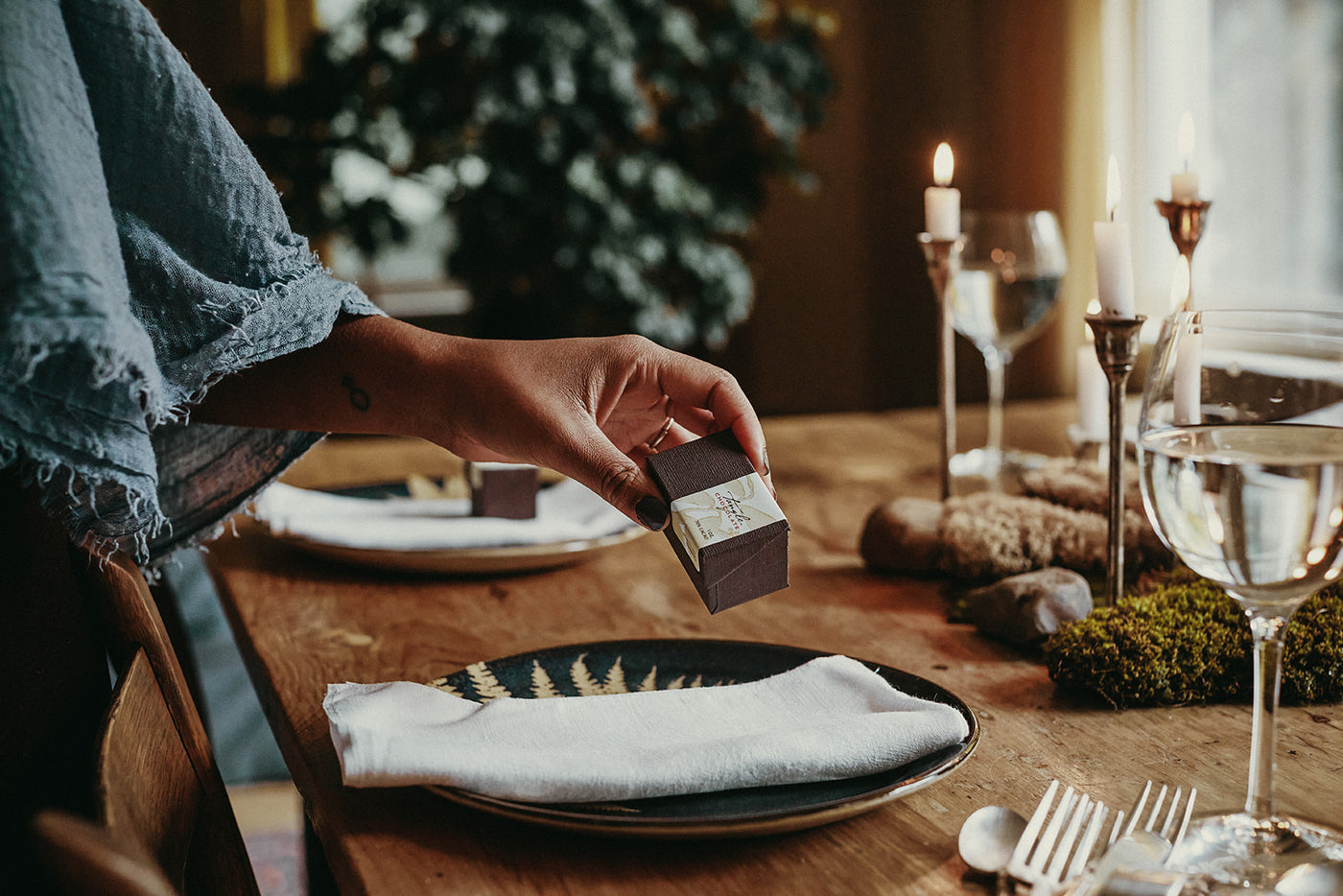 A Black woman's hand and arm holding a small box of Tangle Chocolate THINS, placing the box on a napkin as part of a beautiful table setting with candles, wood table, moss centerpiece.