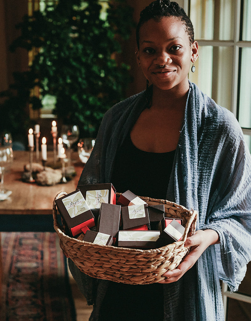 Beautiful Black woman smiling with blue shawl holding a basket filled with boxes of Tangle Chocolate, standing in front of a beautifully set dining room table with candles, warm colors, wood, and green plant in the background. 
