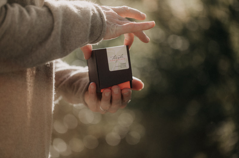 Hands holding a box of Tangle Chocolate THINS, brown box with white label and leaf pattern, coral accent, as though she's about to open the box. Outside setting, wearing a tan cashmere bathrobe.