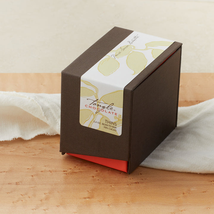 Large closed brown box with a coral accent and white, green, and gold label with leaf pattern that says Tangle Chocolate. on a maple table with a white cloth draped around it. 