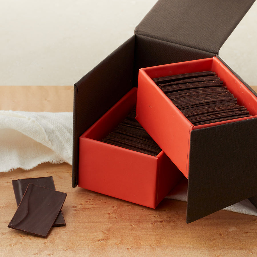 Large open brown with dramatic coral inside with two layers of dark chocolate slivers, and two chocolate slivers lying next to the box. On a maple table with a white cloth draped around it.