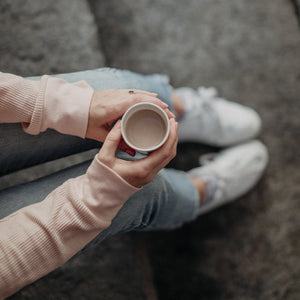 Photo taken from above looking down into a white mug of hot chocolate. Displaying only arms/hands holding the mug, which rests on legs in blue jeans and white sneakers, sitting on stone steps.
