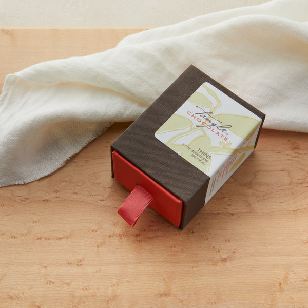 Medium closed brown and coral box with a white, green and gold label with leaf pattern and words "Tangle Chocolate". Coral ribbon to pull open box drawer. The box is filled with slivers of dark chocolate with a couple of the slive On a maple table with a white cloth draped behind it.