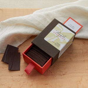 Medium open brown and coral box with a white, green and gold label with leaf pattern and words "Tangle Chocolate". Box drawer is open and a card has popped out the top that says, "You are adorable!" The box is filled with slivers of dark chocolate with a couple of the slivers to the side of the box. On a maple table with a white cloth draped behind it.