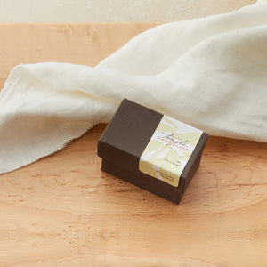 Small closed brown box with white and green label with leaves that says Tangle Chocolate. On a maple table with a white cloth draped behind it.