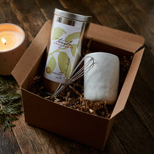 A brown gift box containing a white, handmade mug, small stainless steel whisk, and a 9 oz silver tin of shaved dark chocolate for making hot chocolate.